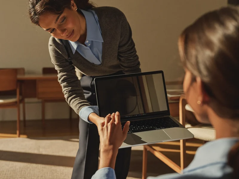 Instructor lângă un cursant așezat la laptop, într-o sală de curs liniștită
